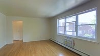 This single-floor space offers a straightforward and functional layout, designed with simplicity and practicality in mind. The main living area is finished with warm-toned hardwood flooring that extends throughout, lending a clean and welcoming feel to the room. Smooth, white walls and a bright white ceiling create an open, airy atmosphere that allows natural light to play a central role in the ambiance of the space.

A prominent window along one wall admits generous natural light and provides views of the surrounding neighborhood, including glimpses of adjacent brick buildings and greenery. The window is paired with a wall-mounted air conditioning unit, ensuring comfort during warmer months. A baseboard heating unit runs along the base of the wall beneath the window, offering efficient warmth throughout the cooler seasons.

The layout is open and unobstructed, offering flexibility in how the space can be arranged and utilized. A doorway visible along one wall connects to additional areas of the unit, suggesting a practical flow between spaces. Overall, the space presents a neat, well-maintained environment with essential amenities in place, making it a functional and comfortable living area.