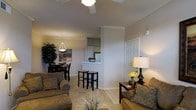 This single-floor residence is designed with an open-concept layout that seamlessly connects the living, dining, and kitchen areas into one cohesive living space. Crown molding runs throughout the space, adding a classic architectural touch that complements the neutral, warm-toned walls.

The living area is generously proportioned, anchored by a ceiling fan that provides both function and a sense of vertical space. Natural light filters into the room, illuminating the light-toned tile flooring that extends throughout the main areas. A side table with a warm-glowing lamp and decorative yellow flowers adds a welcoming ambiance near the entryway, which features a classic paneled door.

A partial wall with a pass-through counter defines the kitchen area, which includes white cabinetry and a raised breakfast bar accompanied by two bar stools. The kitchen's layout allows for easy interaction with the adjacent living and dining spaces.

The dining area is positioned near a large indoor plant, with a pendant light fixture hanging above the dining table and chairs, creating an intimate setting. Overall, the space presents a comfortable, well-appointed interior with cohesive design elements throughout.