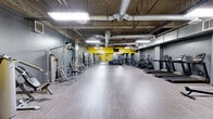 Located at 3075 Wilshire Blvd in Los Angeles, this fitness center offers a generously sized, single-floor layout designed to accommodate a full range of exercise activities. The main exercise room is the centerpiece of the facility, featuring an industrial-style aesthetic with exposed concrete ceilings, visible ductwork, and bright fluorescent lighting that creates a functional, energetic atmosphere. Bold yellow accent walls contrast with the cool gray tones throughout, adding a dynamic visual energy to the space. Light wood-style flooring spans the open floor area, providing a clean and durable surface for workouts. The room is comprehensively equipped with cardio machines including treadmills, stationary bikes, and ellipticals, as well as a wide selection of strength training equipment such as cable machines, weight racks, and multi-use resistance stations. Full-length mirrors along the walls enhance the sense of space and support proper form during training. Three connecting hallways provide organized circulation throughout the facility. The two bathrooms are finished with white subway tile wainscoting, large-format floor tiles, full-length mirrors, and clean gray accents, offering a functional and well-maintained space for post-workout refresh. Overall, the facility presents a practical, well-equipped environment suited for a diverse range of fitness needs.