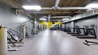 Located at 3075 Wilshire Blvd in Los Angeles, this fitness center offers a generously sized, single-floor layout designed to accommodate a full range of exercise activities. The main exercise room is the centerpiece of the facility, featuring an industrial-style aesthetic with exposed concrete ceilings, visible ductwork, and bright fluorescent lighting that creates a functional, energetic atmosphere. Bold yellow accent walls contrast with the cool gray tones throughout, adding a dynamic visual energy to the space. Light wood-style flooring spans the open floor area, providing a clean and durable surface for workouts. The room is comprehensively equipped with cardio machines including treadmills, stationary bikes, and ellipticals, as well as a wide selection of strength training equipment such as cable machines, weight racks, and multi-use resistance stations. Full-length mirrors along the walls enhance the sense of space and support proper form during training. Three connecting hallways provide organized circulation throughout the facility. The two bathrooms are finished with white subway tile wainscoting, large-format floor tiles, full-length mirrors, and clean gray accents, offering a functional and well-maintained space for post-workout refresh. Overall, the facility presents a practical, well-equipped environment suited for a diverse range of fitness needs.