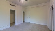 This single-floor space presents a clean, understated aesthetic defined by crisp white walls and trim work that lends a classic character to the interior. The main room is generously proportioned, offering ample open floor space covered in neutral-toned carpet that contributes to a quiet, comfortable atmosphere. Architectural detailing is evident in the paneled wall moldings and substantial baseboards, adding a touch of traditional craftsmanship throughout.

A ceiling fan is centrally positioned overhead, providing both air circulation and a functional focal point for the room. Storage is well-accommodated through a built-in closet with visible shelving and hanging space, alongside an additional door that may lead to further storage or an adjoining room. A separate doorway opens to another area of the unit, suggesting a practical, connected layout.

The space benefits from a ventilation grille integrated into the ceiling, indicating a climate control system. The overall ambiance is bright and neutral, making the space highly versatile and move-in ready for a variety of uses or furnishing styles.