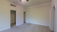 This single-floor space presents a clean, understated aesthetic defined by crisp white walls and trim work that lends a classic character to the interior. The main room is generously proportioned, offering ample open floor space covered in neutral-toned carpet that contributes to a quiet, comfortable atmosphere. Architectural detailing is evident in the paneled wall moldings and substantial baseboards, adding a touch of traditional craftsmanship throughout.

A ceiling fan is centrally positioned overhead, providing both air circulation and a functional focal point for the room. Storage is well-accommodated through a built-in closet with visible shelving and hanging space, alongside an additional door that may lead to further storage or an adjoining room. A separate doorway opens to another area of the unit, suggesting a practical, connected layout.

The space benefits from a ventilation grille integrated into the ceiling, indicating a climate control system. The overall ambiance is bright and neutral, making the space highly versatile and move-in ready for a variety of uses or furnishing styles.