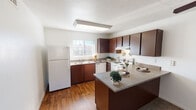 This single-floor residence offers a practical and comfortable layout designed for everyday living. The kitchen is a standout feature, equipped with rich dark-brown cabinetry, granite-style countertops, white appliances including a refrigerator, electric range, and dishwasher, and a peninsula that provides additional prep and dining space. Warm hardwood-style flooring runs throughout the kitchen, creating a clean and inviting atmosphere. Natural light filters in through vertical-blind windows, brightening the space throughout the day.

The open living area flows seamlessly from the kitchen, featuring neutral carpet, a ceiling fan with light fixture, and a textured ceiling. The layout provides generous room for flexible furniture arrangements. The bedrooms continue the carpeted flooring and are each equipped with ceiling fans and natural-light windows. Closet space is included, offering practical storage solutions.

The bathroom is thoughtfully designed with dark wood cabinetry, granite-style countertops, a wide vanity mirror with overhead lighting, and hardwood-style floors. A separate water closet houses the toilet and a full tub and shower combination. The overall aesthetic throughout is cohesive, with consistent dark cabinetry, neutral walls, and warm flooring tones tying each space together.