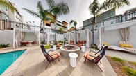 Located at 101 Chamomile Glen in Escondido, California, this single-level outdoor amenity space delivers a resort-inspired experience designed for relaxation and entertainment. At the heart of the space is a generously sized swimming pool with crystal-clear turquoise water, framed by a wide, light-toned tile deck that invites lounging and socializing. A large-format outdoor screen positioned poolside offers an immersive viewing experience, perfect for watching live sports or movies under the open sky.

Surrounding the pool, a series of elegant white-draped private cabanas provide shaded retreats, each furnished with comfortable seating for intimate gatherings. Tall palm trees are thoughtfully interspersed throughout the space, lending a tropical ambiance and natural shade. A central fire pit lounge area features a substantial round concrete fire feature flanked by stylish woven chairs with warm orange cushions and modern side tables, creating an inviting social hub. Additional lounge chairs and market umbrellas line the perimeter, offering ample sun-bathing and relaxation options. The overall design balances contemporary aesthetic with comfortable, functional outdoor living in Southern California's welcoming climate.