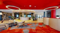 Located at 200 Daytona Blvd in Daytona Beach, Florida, this expansive single-floor space makes an immediate impression with its striking contemporary design. The main lobby area features a bold red ceiling accented by oversized circular pendant lights, paired with a vibrant geometric-patterned carpet in orange, red, and grey tones that anchors the welcoming entrance. A curved, wood-paneled front desk with dual service stations sits beneath a matching red ceiling detail, framed by large windows that invite natural light and outdoor views.

At the heart of the space is a curved bar and lounge area finished in warm natural wood, surrounded by bar-height seating and vivid yellow patterned flooring — a lively social hub ideal for gathering. Adjacent lounge zones offer a variety of seating arrangements, from plush armchairs to communal tables, all set against light hardwood flooring and softly curved architectural wall panels with integrated cove lighting.

A spacious amenity lounge opens toward floor-to-ceiling glazed doors leading to a landscaped outdoor area, with a foosball table and casual seating creating a relaxed, recreational atmosphere. An elevator lobby showcases curated wall art and geometric LED ceiling detailing. Throughout, lush indoor plants, cylindrical columns in slate blue, and a cohesive mix of bold and neutral tones give the space an energetic yet refined character.