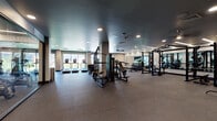 This contemporary fitness center is designed to accommodate a wide range of workout styles within a thoughtfully organized, single-floor layout. The space is finished with durable rubber flooring throughout, dark ceilings with recessed and linear LED lighting, and floor-to-ceiling mirrors that enhance the sense of openness and allow users to monitor their form.

The cardio zone is positioned along a bank of large windows, offering natural light and outdoor views while using treadmills, upright and recumbent bikes, and cycling machines. A dedicated strength training area houses Matrix power racks, cable machines, multi-functional cable stations, and a full dumbbell rack with an integrated mirror wall, providing comprehensive options for resistance training.

A separate functional training room features a modular rig equipped with TRX suspension trainers, kettlebells, medicine balls, and a heavy punching bag, making it ideal for high-intensity and cross-training workouts. Additional built-in wooden cubbies offer convenient storage for personal items. The facility is accessed through sleek glass-paneled doors and benefits from a ceiling fan for added air circulation. The overall ambiance is clean, professional, and purposefully designed to support a full-spectrum fitness experience.