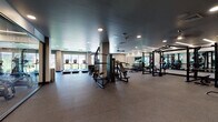 This contemporary fitness center is designed to accommodate a wide range of workout styles within a thoughtfully organized, single-floor layout. The space is finished with durable rubber flooring throughout, dark ceilings with recessed and linear LED lighting, and floor-to-ceiling mirrors that enhance the sense of openness and allow users to monitor their form.

The cardio zone is positioned along a bank of large windows, offering natural light and outdoor views while using treadmills, upright and recumbent bikes, and cycling machines. A dedicated strength training area houses Matrix power racks, cable machines, multi-functional cable stations, and a full dumbbell rack with an integrated mirror wall, providing comprehensive options for resistance training.

A separate functional training room features a modular rig equipped with TRX suspension trainers, kettlebells, medicine balls, and a heavy punching bag, making it ideal for high-intensity and cross-training workouts. Additional built-in wooden cubbies offer convenient storage for personal items. The facility is accessed through sleek glass-paneled doors and benefits from a ceiling fan for added air circulation. The overall ambiance is clean, professional, and purposefully designed to support a full-spectrum fitness experience.
