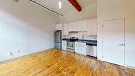 Located at 95 Chestnut St in Providence, RI, this one-bedroom, one-bathroom loft-style unit showcases the character of a thoughtfully converted historic building. The open layout is anchored by warm, original hardwood floors that run throughout the main living areas, complemented by soaring ceilings that lend an airy, expansive feel to the compact yet efficient floor plan. Exposed wooden beams and a striking structural timber post add authentic industrial charm, while a prominent exposed brick wall with large, black-framed windows floods the bedroom with natural light and urban views. The kitchen is well-appointed with white shaker cabinetry extending to ceiling height, dark countertops, and a full suite of stainless steel appliances including a refrigerator, electric range, dishwasher, and sink. Exposed ductwork and pendant lighting contribute to the loft's industrial aesthetic. The bathroom is clean and functional, finished with light tile flooring, a vanity with cabinet storage, a mirror, and a tiled shower surround. A hallway connects the rooms efficiently, completing this well-organized, character-rich urban residence in the heart of Providence.