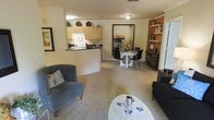 This single-floor residence showcases an open-concept layout that integrates the living, dining, and kitchen areas into one cohesive, flowing space. The living area is anchored by plush carpeting in warm beige tones, complemented by comfortable seating including a cushioned accent chair and a sleek sofa arrangement around a round white coffee table. A framed piece of wall art adds a decorative touch to the space.

The kitchen features light maple cabinetry with dark countertops and is defined by a wraparound breakfast bar that provides both a functional workspace and a natural visual boundary between the cooking and living areas. Appliances are neatly integrated beneath the upper cabinetry, and a pass-through design keeps the space feeling open and connected.

Adjacent to the kitchen, the dining area accommodates a round white table surrounded by contemporary black chairs, making it ideal for everyday dining. A bold abstract artwork serves as a striking focal point on the wall nearby. To the right, built-in shelving with an accent wall in warm terracotta tones provides decorative display space, adding depth and character to the overall ambiance.