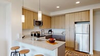 This thoughtfully designed one-bedroom, one-bathroom unit offers a contemporary living experience across a single floor. The open-concept kitchen and living area serves as the heart of the unit, featuring a kitchen outfitted with light wood upper cabinetry, contrasting dark lower cabinets, granite countertops, and a full suite of stainless steel appliances including a range, microwave, dishwasher, and refrigerator. A peninsula with bar seating and stylish pendant lighting provides both prep space and casual dining convenience. Light hardwood-style flooring flows throughout, enhancing the bright, airy atmosphere. The living area is anchored by expansive floor-to-ceiling picture windows that frame impressive views of the surrounding cityscape and mountain range beyond, flooding the space with natural light. The bedroom is a calm, well-proportioned retreat with its own large windows offering the same scenic vistas, a ceiling fan, and access to a dedicated closet. The bathroom features large-format tile flooring, clean white walls, and a wood-accented vanity for a modern, polished look. A hallway connects the main living spaces efficiently. The unit's clean lines, neutral palette, and abundant natural light give it a fresh, modern character throughout.