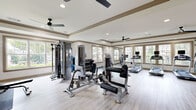 Located at 1110 Ballpark Ln in Lawrenceville, GA, this fitness center offers a well-equipped, single-floor exercise room designed to accommodate a full range of workout routines. The space features elegant tray ceilings with crown molding trim details and recessed lighting, complemented by dual ceiling fans that promote airflow throughout. Light wood-look flooring runs across the entire floor, lending a clean, modern aesthetic.

Large framed windows line the walls, flooding the room with natural light and offering views of the surrounding greenery. Full-length mirrors are strategically positioned to visually expand the space and assist users during workouts. The cardio section is well-stocked with multiple treadmills and elliptical machines, each equipped with individual display screens. A dedicated strength training area houses a multi-station cable machine, adjustable benches, a power tower, and a full rack of dumbbells. Exercise balls and floor mats provide space for functional and flexibility training.

A wall-mounted television, fire safety equipment, and clearly marked exits reflect a commitment to safety and convenience. The overall ambiance is bright, open, and inviting, making this fitness center a comprehensive and comfortable environment for residents and users seeking an effective workout experience.