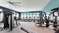 Located at 7600 Heron Cir in Vero Beach, Florida, this single-story fitness center offers a generously sized, thoughtfully designed workout facility spread across two distinct areas. The space is defined by its vibrant teal accent walls, crisp white ceilings, and neutral-toned carpet flooring, creating an energetic yet clean aesthetic throughout.

The main workout area is expansive and bathed in natural light from a row of large windows along the exterior wall, offering views of the surrounding Florida landscape. This section is equipped with multiple treadmills, elliptical machines, a multi-station cable and strength system, a dumbbell rack, adjustable benches, and a wall-mounted television — all anchored beneath a large ceiling fan that promotes air circulation. Full-length mirrors line one wall, enhancing the sense of space.

A second dedicated room features lighter hardwood-style flooring and vaulted ceilings with high clerestory windows that flood the area with natural light. This space is outfitted for functional and cross-training, including Peloton-style spin bikes, a heavy punching bag, medicine balls, battle ropes, plyo boxes, and additional strength accessories. A large mirror and ceiling fan complete the room.

The facility also includes a transitional area with sliding barn doors, built-in shelving, and access to an ancillary room, providing versatility for various fitness programming needs.