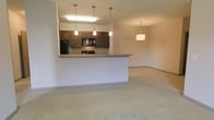 This single-floor space is designed with an open-concept layout that seamlessly connects the kitchen and living area, creating a cohesive and functional environment. The kitchen is a standout feature, equipped with rich dark wood cabinetry that provides ample storage, black appliances including a refrigerator, microwave, and range, and a sleek countertop peninsula that doubles as a breakfast bar. Three pendant lights hang above the kitchen workspace, providing focused task lighting while adding a contemporary decorative touch. An additional pendant light fixture illuminates the adjoining living area, contributing to a warm and welcoming ambiance throughout the space. The living area is finished with neutral wall-to-wall carpeting that adds comfort underfoot and complements the overall muted, warm color palette of the interior. Recessed lighting in the kitchen ceiling supplements the pendant fixtures for well-distributed illumination. The open sightlines between the kitchen and living room make the space feel expansive and airy, ideal for both everyday living and entertaining. The consistent neutral tones across walls, flooring, and trim create a clean, move-in-ready aesthetic that offers a versatile backdrop for a variety of interior design styles.