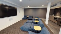 This single-floor space is designed as a casual lounge or media room, offering a comfortable and inviting environment for relaxation and group gatherings. The room is anchored by a large wall-mounted flat-screen display, making it well-suited for screenings, presentations, or casual viewing sessions.

The seating arrangement is a standout feature, comprising an array of deep, modular low-profile sofas and chairs upholstered in a rich navy blue fabric. The plush, cushioned seating is arranged in a tiered, informal layout to maximize sightlines toward the screen. Compact round side tables with light wood tops are interspersed throughout the seating area, providing convenient surfaces for drinks or personal items.

The flooring consists of a textured, medium-toned carpet that adds warmth and acoustic comfort to the room. The walls are finished in a two-tone palette — crisp white on the upper portions and a bold charcoal grey on the accent wall behind the seating — creating a modern, cinema-inspired aesthetic. Recessed ceiling lighting provides even, ambient illumination throughout the space. The overall design balances functionality and comfort, making this lounge an ideal communal space for entertainment and social interaction.