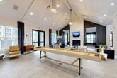 Located in Katy, TX, this expansive single-floor community clubhouse offers a well-rounded amenity experience across a generously sized footprint. The main lounge is an architectural highlight, featuring soaring vaulted ceilings with exposed white-beamed details, elegant pendant lighting, and large black-framed glass doors that invite abundant natural light. A dramatic fireplace anchors the seating area, framed by a bold dark accent wall with textured geometric paneling and a wall-mounted TV, creating a sophisticated gathering space. A full-length shuffleboard table serves as a central focal point on the light tile flooring.

Adjacent to the lounge, a dedicated games room features an air hockey table, dark accent walls, crown molding, and large windows with dramatic floor-length drapes. A business center area offers built-in desk workstations beneath pendant lighting, accompanied by a wall-mounted display and a textured accent wall.

The fitness center is bright and airy, with high ceilings, a ceiling fan, and wide windows overlooking the outdoor pool area. It is equipped with cardio machines, free weights, kettlebells, a multi-station cable machine, and floor-to-ceiling mirrors. A well-appointed kitchen features dark cabinetry, white countertops, a stainless refrigerator, and a textured mosaic accent wall. A dedicated package room with metal shelving completes this comprehensive community facility.