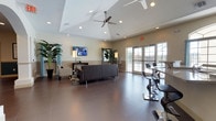 This single-floor communal space offers a well-appointed and inviting atmosphere designed for both relaxation and casual interaction. The lounge is anchored by soaring vaulted ceilings with a recessed tray detail and a modern ceiling fan, lending the room a sense of openness and airiness. Recessed lighting throughout ensures the space is well-lit at all hours.

The flooring consists of large-format dark tile, providing a sleek, contemporary foundation that contrasts beautifully with the lighter walls and ceiling. The seating area features a dark leather sofa and an accent chair with a patterned upholstery, arranged around a wall-mounted flat-screen television — creating a comfortable social hub. Tall indoor plants are strategically placed throughout, adding a refreshing natural element to the modern interior.

A high-top bar table with modern bar stools is positioned near a wall of French doors, offering a casual dining or gathering spot bathed in natural light. The French doors open to an outdoor area, seamlessly connecting the interior to the exterior environment. Warm-toned wall sconces complement the neutral color palette, completing the polished yet welcoming ambiance of this thoughtfully designed communal lounge.