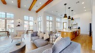 This single-story space is designed with an open-concept layout that seamlessly connects a spacious lounge, dining area, and fully equipped kitchen. The architecture is defined by dramatically high ceilings adorned with rich, exposed wood beams that add warmth and character against crisp white walls. Light hardwood flooring runs throughout, unifying the expansive floor plan.

The lounge area is anchored by a floor-to-ceiling limestone fireplace with a linear gas insert, topped by a wall-mounted television — serving as a striking focal point. Comfortable gray upholstered sofas and seating arrangements complete the area.

The kitchen is outfitted with warm, natural wood cabinetry featuring matte black hardware, light quartz countertops, a large center island with open shelving, stainless steel appliances, and a statement range hood. Open floating shelves provide additional display and storage space.

The dining area benefits from towering, multi-pane windows that flood the space with natural light and offer views of the surrounding outdoor courtyard and grounds. Wall sconces and pendant lighting throughout contribute to a layered, inviting ambiance.

The entry features wood-framed glass double doors, transom windows above, and additional seating, creating a welcoming first impression consistent with the space's modern farmhouse aesthetic.