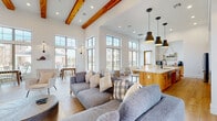 This single-story space is designed with an open-concept layout that seamlessly connects a spacious lounge, dining area, and fully equipped kitchen. The architecture is defined by dramatically high ceilings adorned with rich, exposed wood beams that add warmth and character against crisp white walls. Light hardwood flooring runs throughout, unifying the expansive floor plan.

The lounge area is anchored by a floor-to-ceiling limestone fireplace with a linear gas insert, topped by a wall-mounted television — serving as a striking focal point. Comfortable gray upholstered sofas and seating arrangements complete the area.

The kitchen is outfitted with warm, natural wood cabinetry featuring matte black hardware, light quartz countertops, a large center island with open shelving, stainless steel appliances, and a statement range hood. Open floating shelves provide additional display and storage space.

The dining area benefits from towering, multi-pane windows that flood the space with natural light and offer views of the surrounding outdoor courtyard and grounds. Wall sconces and pendant lighting throughout contribute to a layered, inviting ambiance.

The entry features wood-framed glass double doors, transom windows above, and additional seating, creating a welcoming first impression consistent with the space's modern farmhouse aesthetic.