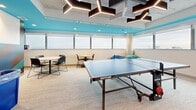 Located on Bay Street South in Hamilton, Ontario, this energetic recreation room is designed to offer a dynamic and engaging social environment. The space is defined by its eye-catching ceiling installation, which features geometric, hexagonal acoustic panels in dark tones accented by angular LED light strips — creating a bold, industrial-modern aesthetic against the exposed concrete structure above.

The room is equipped with a Kettler Berlin Pro Outdoor ping pong table and a foosball table, providing ample entertainment options. Comfortable bean bag chairs in blue and green are arranged near the windows, offering a more casual lounging area. Small round tables with modern chairs are positioned along the window wall, ideal for informal gatherings or casual work sessions.

A continuous band of horizontal windows wraps along two walls, flooding the room with natural light and offering sweeping views of the Hamilton city skyline. A vivid turquoise ceiling section adds a bold pop of colour, complemented by a striking diagonal geometric mural in teal, brown, and white tones on one accent wall. Neutral-toned carpet underfoot and clean white walls balance the room's more vibrant design elements. Recycling and waste stations are conveniently integrated into the space, reflecting a commitment to sustainability.