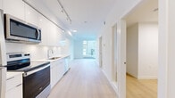 This contemporary single-floor unit offers a clean, minimalist aesthetic defined by an all-white palette, light hardwood flooring, and an abundance of natural light. The open-concept living and kitchen area forms the heart of the space, where a streamlined galley-style kitchen runs along one wall, outfitted with floor-to-ceiling white cabinetry, white quartz countertops, a stainless steel undermount sink, and a full suite of stainless steel appliances including a range, over-the-range microwave, dishwasher, and refrigerator. Track lighting overhead provides functional illumination throughout. The living area extends toward a wall of floor-to-ceiling glazing and sliding doors that open to a private balcony, offering views of the surrounding contemporary building complex and distant mountains beyond. A dedicated bedroom features a large casement window that continues the theme of natural light and outdoor connection. The bathroom is neatly appointed with a soaker tub and shower combination, white tile surround, and matching clean-line fixtures. A hallway connects all areas of the unit efficiently, with additional doors providing access to in-suite storage. The overall ambiance is bright, fresh, and well-organized, reflecting modern residential design standards.