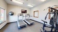 Located at 5617 Muirfield Dr SW in Cedar Rapids, Iowa, this compact fitness center offers a well-equipped workout space designed for convenient, everyday exercise. The room is finished with durable low-pile carpet flooring and neutral light-colored walls, creating a clean and functional environment. Fluorescent ceiling fixtures provide consistent, even lighting throughout the space.

The fitness center is equipped with two side-by-side treadmills positioned toward one wall, a multi-function cable weight machine suitable for a variety of strength training exercises, and additional fitness accessories including a stability ball and a flat workout bench. A wall-mounted flat-screen television is positioned above the treadmills, allowing users to stay entertained during cardio sessions.

Multiple windows with warm wood trim line the walls and allow natural light to enter the room. Notably, windows on one side offer a direct view of an outdoor pool area, adding a pleasant visual element to workouts. A glass-paned French door with matching wood trim provides access to and from the space, contributing a polished, classic aesthetic. The room also includes a hand sanitizer dispenser and fire safety features, reflecting attention to both hygiene and safety standards.