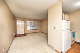 This single-floor unit offers a practical and straightforward layout designed for comfortable everyday living. The living area is a notable feature of the space, finished with light-toned wall-to-wall carpeting and neutral beige walls that create a clean, understated atmosphere. Large windows allow generous natural light to fill the room, and a front entry door with an adjacent window panel provides a welcoming point of arrival. A ceiling fan is present overhead, adding functional comfort. Textured popcorn ceilings are consistent throughout the unit.

The galley-style kitchen is efficiently arranged, with white cabinetry lining both sides, a white electric range, refrigerator, and a stainless steel sink. Wood-look flooring runs through the kitchen, contrasting neatly with the carpeted living spaces. Upper cabinets provide ample storage, and a door at the end of the kitchen connects to an adjacent area of the unit.

The bathroom is well-appointed, featuring a single-sink vanity with a large mirror, wood-toned cabinetry below, a toilet, and a full bathtub with a shower curtain rod. Wood-look flooring continues here, maintaining visual consistency. The overall ambiance of the unit is neutral, functional, and move-in ready.