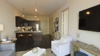 This single-floor residence offers a thoughtfully designed open-concept layout that connects the kitchen and living areas in a seamless, functional arrangement. The kitchen is a standout feature, showcasing rich dark wood cabinetry, stainless steel appliances, and a breakfast bar with bar-stool seating — ideal for casual dining or entertaining. A cluster of Edison-style pendant lights hangs above the kitchen island, casting a warm, inviting glow throughout the space. Dark hardwood flooring runs throughout, grounding the interior with a sense of depth and sophistication. The living area transitions naturally from the kitchen and is styled with a crisp white accent chair, a glass-top coffee table, and tasteful decorative elements that contribute to the space's warm, curated ambiance. Neutral wall tones provide a clean backdrop that complements the darker design elements and enhances the brightness of the room. A hallway leading to additional rooms is visible in the background, suggesting a practical and well-organized floor plan. Overall, the space strikes a balance between modern design and comfortable living, making it both visually appealing and highly functional.