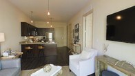This single-floor residence offers a thoughtfully designed open-concept layout that connects the kitchen and living areas in a seamless, functional arrangement. The kitchen is a standout feature, showcasing rich dark wood cabinetry, stainless steel appliances, and a breakfast bar with bar-stool seating — ideal for casual dining or entertaining. A cluster of Edison-style pendant lights hangs above the kitchen island, casting a warm, inviting glow throughout the space. Dark hardwood flooring runs throughout, grounding the interior with a sense of depth and sophistication. The living area transitions naturally from the kitchen and is styled with a crisp white accent chair, a glass-top coffee table, and tasteful decorative elements that contribute to the space's warm, curated ambiance. Neutral wall tones provide a clean backdrop that complements the darker design elements and enhances the brightness of the room. A hallway leading to additional rooms is visible in the background, suggesting a practical and well-organized floor plan. Overall, the space strikes a balance between modern design and comfortable living, making it both visually appealing and highly functional.