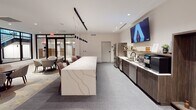 Located at 703 E Main St in the heart of Richmond, Virginia, this single-floor amenity space combines contemporary design with a versatile range of social and recreational features. The entrance area welcomes visitors with large black-framed glass partitions, offering views of a stairwell and an adjacent meeting area furnished with a round wood table and patterned upholstered chairs — an inviting setting for casual conversations or small gatherings.

The central lounge and kitchenette area serves as the hub of the space. A long, dramatically veined quartz island anchors the room, illuminated by sleek pendant lighting above. Dark wood cabinetry lines one wall, housing a stainless steel refrigerator, sink, coffee station, and integrated appliances. A wall-mounted flat-screen TV completes the kitchen bar setup, making it ideal for both everyday use and social events.

Beyond the kitchen, the space opens into a generously sized recreation and lounge area featuring a pool table, multiple seating arrangements including high-top bistro tables with vibrant blue and neutral upholstered chairs, and additional lounge zones. A dedicated media lounge with a plush green sectional sofa, herringbone-patterned accent wall, warm wood paneling, and a mounted flat-screen TV creates an intimate retreat within the larger space. Recessed lighting and neutral carpeting throughout contribute to the polished, welcoming atmosphere.