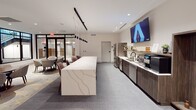 Located at 703 E Main St in the heart of Richmond, Virginia, this single-floor amenity space combines contemporary design with a versatile range of social and recreational features. The entrance area welcomes visitors with large black-framed glass partitions, offering views of a stairwell and an adjacent meeting area furnished with a round wood table and patterned upholstered chairs — an inviting setting for casual conversations or small gatherings.

The central lounge and kitchenette area serves as the hub of the space. A long, dramatically veined quartz island anchors the room, illuminated by sleek pendant lighting above. Dark wood cabinetry lines one wall, housing a stainless steel refrigerator, sink, coffee station, and integrated appliances. A wall-mounted flat-screen TV completes the kitchen bar setup, making it ideal for both everyday use and social events.

Beyond the kitchen, the space opens into a generously sized recreation and lounge area featuring a pool table, multiple seating arrangements including high-top bistro tables with vibrant blue and neutral upholstered chairs, and additional lounge zones. A dedicated media lounge with a plush green sectional sofa, herringbone-patterned accent wall, warm wood paneling, and a mounted flat-screen TV creates an intimate retreat within the larger space. Recessed lighting and neutral carpeting throughout contribute to the polished, welcoming atmosphere.