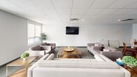 Located at 1355 Commissioners Rd W in London, Ontario, this single-floor communal lounge offers a thoughtfully designed gathering space with a clean, modern aesthetic. The open-concept layout is organized into several functional zones, seamlessly blending lounge and dining areas within a comfortable footprint.

The seating area anchors one side of the room, furnished with a large white sofa, tufted armchairs in soft mauve tones, and a warm-toned wooden coffee table — all centered beneath a wall-mounted flat-screen television. Natural light filters through multiple windows fitted with roller blinds, creating a bright and welcoming atmosphere. Decorative potted plants add a touch of greenery throughout the space.

A round glass-top dining table with wooden chairs provides an intimate setting for smaller gatherings, while a larger rectangular dining table nearby accommodates additional guests. A floor-to-ceiling glass partition wall elegantly separates a semi-private area from the main lounge, maintaining visual openness while offering a sense of enclosure.

Underfoot, a neutral patterned carpet runs throughout, complementing the crisp white walls and drop-tile ceiling with recessed lighting. Access doors and built-in shelving along the perimeter add practical functionality. The overall ambiance is polished, comfortable, and well-suited for both casual socializing and organized gatherings.