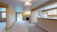 This single-floor residence offers a functional and comfortable layout with a warm, neutral color palette throughout. The living area serves as the centerpiece of the space, featuring plush wall-to-wall carpeting, a ceiling fan with integrated lighting, and a classic white fireplace that adds a focal point to the room. A full-length sliding glass door dressed with vertical blinds fills the living area with natural light and provides direct views of the greenery outside.

The kitchen is positioned adjacent to the living area in an open-concept arrangement, separated by a half-wall counter that allows visual connectivity between the two spaces. The kitchen is equipped with ample white cabinetry trimmed in warm wood tones, light-colored countertops, a white appliance suite including a refrigerator, electric range, and dishwasher, and durable wood-look flooring.

The entry foyer features hard flooring and provides a clear sightline into the main living space. A bedroom offers carpeted flooring, a window with outdoor views, and a baseboard heater. The bathroom includes a full tub-shower combination with clean white surroundings and wood-look flooring. Track lighting accents are present in select areas, adding functional illumination throughout the space.