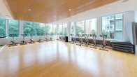 Located at 800 P Street NW in Washington, D.C., this contemporary fitness studio is designed to deliver an energizing and functional workout environment. The single-floor space is anchored by a generously sized exercise room outfitted with a full row of stationary cycling bikes arranged along expansive floor-to-ceiling windows that flood the interior with abundant natural light and frame views of the surrounding D.C. neighborhood. The warm bamboo hardwood flooring spans the entire space, providing both aesthetic appeal and a practical surface suitable for a range of fitness activities. A warm wood-paneled ceiling with recessed lighting adds an inviting, refined quality to the room, complementing the clean white walls throughout. Full-length mirrors line one wall, visually expanding the space and supporting group exercise and barre classes, with a mounted ballet barre visible along the mirrored section. Wall-mounted flat-screen televisions are positioned for easy viewing during workouts. Motivational typography displayed along the windows adds an energetic, purposeful character to the room. The studio's open layout, combined with its bright, airy atmosphere and thoughtful design details, creates a welcoming and high-functioning environment well-suited for group fitness classes and individual training sessions.