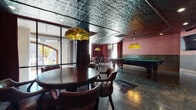 This single-floor entertainment space exudes a classic, upscale lounge atmosphere through a carefully curated combination of vintage and industrial design elements. The standout architectural feature is the ornate pressed-tin ceiling, finished in a metallic silver tone that adds texture and visual depth throughout the space. Polished concrete floors reflect ambient light, lending an industrial edge that contrasts elegantly with the warmer decorative accents.

The main area is anchored by two full-size pool tables with green felt surfaces, each illuminated by Tiffany-style stained-glass pendant lights that cast warm, colorful tones. Surrounding the tables, richly upholstered leather game chairs and round wooden tables provide comfortable seating for card games or casual gatherings. Deep red curtains and rose-toned accent walls contribute to the room's intimate, lounge-like ambiance.

Large glass walls and windows along one side allow natural light to filter in, offering views of the exterior surroundings. Decorative wood trim and wainscoting frame key transitions within the space, adding refinement. A separate adjacent area with polished concrete flooring and exposed ductwork provides additional flexible space. Cue racks, bar-height seating, and a wall-mounted television round out the functional amenities of this well-appointed entertainment venue.