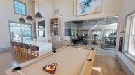 This single-floor clubhouse, known as The Retreat, is thoughtfully designed to offer a well-rounded amenity experience. The main lounge serves as the heart of the space, anchored by a dramatic floor-to-ceiling stacked stone fireplace with a mounted flat-screen television above. Soaring ceilings amplify the sense of openness, while large windows on multiple walls invite generous natural light and views of the surrounding outdoor areas, including a pool visible through one of the exits.

The lounge transitions seamlessly into a game area equipped with a full-size billiards table, complemented by contemporary pendant lighting and abstract artwork overhead. A built-in credenza with open wood shelving provides functional display and storage space, incorporating warm wood tones that balance the room's modern, neutral palette. Bar-height seating along a white counter adds a casual social element to the space.

Adjacent to the lounge, a glass-enclosed fitness center is fully equipped with cardio machines, stationary bikes, and strength training equipment, offering visibility from the main area while maintaining a dedicated workout environment. Warm hardwood flooring runs throughout, contributing to the cohesive and welcoming ambiance of the entire clubhouse.