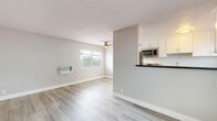 This single-floor unit offers a clean, modern aesthetic throughout, with light wood-look flooring and a neutral gray-and-white color palette that creates a fresh, cohesive atmosphere. The open living area is generously proportioned, filled with natural light from wide, blinds-fitted windows, and includes a ceiling fan for added comfort. A wall-mounted AC unit provides climate control in both the living area and the bedroom. The pass-through kitchen is thoughtfully designed with white shaker-style cabinetry, dark countertops, stainless steel appliances including a gas range and built-in microwave, and a functional layout that keeps the space feeling connected and open. Two well-sized bedrooms each feature light flooring, neutral walls, and windows that frame views of mature palm trees, reinforcing a warm, sunny character. The bathroom is outfitted with a spacious white vanity featuring a dark countertop, ample drawer storage, and a well-lit mirror, alongside a separate tiled tub-and-shower combination. Throughout the unit, clean lines, updated finishes, and abundant natural light combine to create a comfortable and practical living space.