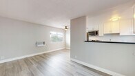 This single-floor unit offers a clean, modern aesthetic throughout, with light wood-look flooring and a neutral gray-and-white color palette that creates a fresh, cohesive atmosphere. The open living area is generously proportioned, filled with natural light from wide, blinds-fitted windows, and includes a ceiling fan for added comfort. A wall-mounted AC unit provides climate control in both the living area and the bedroom. The pass-through kitchen is thoughtfully designed with white shaker-style cabinetry, dark countertops, stainless steel appliances including a gas range and built-in microwave, and a functional layout that keeps the space feeling connected and open. Two well-sized bedrooms each feature light flooring, neutral walls, and windows that frame views of mature palm trees, reinforcing a warm, sunny character. The bathroom is outfitted with a spacious white vanity featuring a dark countertop, ample drawer storage, and a well-lit mirror, alongside a separate tiled tub-and-shower combination. Throughout the unit, clean lines, updated finishes, and abundant natural light combine to create a comfortable and practical living space.