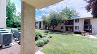 This single-floor residential unit is part of a classic brick multi-building complex surrounded by mature trees, manicured lawns, and well-kept landscaping. The exterior presents a welcoming courtyard-style setting with lush greenery visible from a covered entryway supported by a sturdy brick column, offering a pleasant outdoor perspective under open skies.

Inside, the living area is finished with warm, neutral-toned walls and plush wall-to-wall carpeting that creates a soft, comfortable atmosphere throughout. A standout feature of the main living space is a full-width sliding glass door dressed with vertical blinds, allowing generous natural light to pour in while providing direct access to the outdoors and framing views of the verdant courtyard beyond. A ceiling fan with light fixture adds both function and a finishing touch to the open layout.

The interior flows naturally between the living space and additional rooms, with clean sightlines and an open, airy feel. The rooms are finished consistently with the same neutral palette—warm off-white walls, crisp white baseboards, and carpeted floors—providing a calm, blank-canvas environment that is highly adaptable. The overall ambiance is clean, quiet, and light-filled, making it a comfortable and versatile living space within a peaceful, green residential setting.