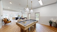 This well-appointed single-floor community space offers an inviting, open-concept layout designed for both relaxation and recreation. The main gathering area is anchored by a classic fireplace with a white tile surround and decorative mantle, complemented by warm hardwood-style flooring that flows seamlessly throughout. Stylish pendant lighting with brass accents illuminates a full-sized billiard table, making the game area a natural focal point of the space.

The lounge area is generously proportioned, featuring a mix of comfortable seating including plush sofas and modern accent chairs arranged around a sculptural geometric coffee table. Crown molding and wainscoting add architectural refinement to the bright white walls, while recessed lighting enhances the airy ambiance.

A well-equipped kitchen runs adjacent to the main area, featuring sleek dark cabinetry, expansive white quartz countertops, stainless steel appliances, and a view of the outdoor pool area through a large window. A dedicated dining nook with a round table and wishbone chairs provides an intimate setting for smaller gatherings.

Multiple glass-paned doors and large windows frame views of lush greenery and the surrounding outdoor areas, bathing the entire space in natural light and creating a seamless indoor-outdoor connection.