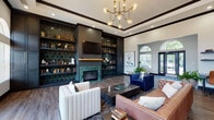 This elegantly designed single-floor communal space blends contemporary sophistication with classic architectural detail. The main lounge area commands attention with soaring tray ceilings trimmed in dark crown molding, a statement brass sputnik chandelier, and an abundance of natural light streaming through a series of arched windows and French doors that open to the outdoors. Rich hardwood-style flooring runs throughout, grounding an eclectic mix of seating that includes a tufted cognac leather sofa, a deep navy velvet sectional, and cream upholstered accent chairs arranged around a substantial coffee table. A dramatic built-in wall unit, finished in dark cabinetry and accented with bold emerald geometric tile, houses a mounted television, a fireplace with a marble surround, and curated open shelving. Adjacent to the main lounge, a dedicated work area features wall-mounted shelving and task seating, ideal for productivity. The entry foyer is bright and welcoming, with arched transom windows, lush potted greenery, and polished flooring that leads seamlessly into the lounge. A compact kitchenette with white quartz countertops, stainless steel appliances, dark subway tile backsplash, and bar-height seating rounds out the space, providing practical amenities in a refined setting.
