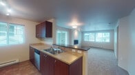 Located at 15100 SW Barrows Rd in Beaverton, Oregon, this single-floor residence offers a thoughtfully designed open-concept layout that seamlessly connects the kitchen and living areas. The kitchen is well-equipped with rich dark wood cabinetry, stainless steel appliances including a refrigerator, range, microwave, and dishwasher, complemented by tile flooring and track lighting. A peninsula with a double basin sink and bar-top overhang provides both functional workspace and an informal dining option, while opening directly to the living area. The spacious living room features soft wall-to-wall carpeting, a wall-mounted fireplace as a focal point, and abundant natural light streaming in through large windows and a sliding glass door that leads to a private balcony with outdoor views. The bedroom is a calm, carpeted retreat with a dedicated window and baseboard heating. The bathroom includes rich wood vanity cabinetry, tile flooring, a large mirror, and an adjacent tub and shower area. A second bathroom offers similar finishes with a single-sink vanity, tile floors, and a toilet. Baseboard heating is present throughout the unit. The overall ambiance is clean, neutral, and move-in ready.