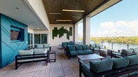 Located along the scenic Boardwalk Trail in Austin, TX, this single-level covered lounge and terrace space offers a sophisticated blend of indoor comfort and outdoor connectivity. The architecture features soaring ceilings clad in warm wood slats, accented by linear LED lighting that enhances the modern aesthetic throughout. A prominent geometric moss wall installation serves as a striking focal point, adding a natural, textural element to the otherwise clean-lined interior.

The space transitions seamlessly between a partially enclosed interior lounge and an open-air terrace, connected via expansive floor-to-ceiling glass doors. The interior lounge area is anchored by teal upholstered sofas arranged around low coffee tables, with a mounted flat-screen television and a bold teal accent wall featuring angular paneling. A separate nook with lighter seating and a second mounted display offers an additional gathering area.

The outdoor terrace extends the lounge's functionality, furnished with coordinated teal cushioned seating and glass-panel railings that preserve unobstructed views of the surrounding waterway and lush tree canopy. A built-in wet bar with counter seating enhances the outdoor entertaining experience. Large-format tile flooring flows consistently throughout, reinforcing the cohesive, upscale ambiance of the entire space.