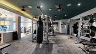 Located in Charlotte, NC, this comprehensive fitness center is thoughtfully designed across a single floor, offering two distinct workout zones that cater to a wide range of training styles and fitness goals. The strength training area features a dramatic teal-painted ceiling accented with recessed lighting and ceiling fans, complemented by patterned carpet flooring. This zone is outfitted with a multi-station cable machine, power racks, weight benches, medicine balls, a heavy punching bag, and floor-to-ceiling mirrors that amplify the sense of space. An adjacent transitional area includes a built-in counter with a beverage refrigerator, exposed brick columns, and elegant pendant lighting, adding a refined touch to the functional environment. The studio and cardio zone offers a bright, airy contrast with light hardwood flooring, exposed wooden ceiling beams, and large windows that invite abundant natural light. This area accommodates spin bikes, a rowing machine, suspension training equipment, wall-mounted storage, and a large display screen for guided workouts. A dedicated cardio room rounds out the facility, housing treadmills, ellipticals, and stair climbers beneath a beamed ceiling with ample natural light from oversized windows. The overall design blends modern fitness functionality with warm, stylish architectural details.
