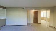 This single-floor space presents an open, interconnected layout that promotes a functional flow between rooms. The main living area features wall-to-wall carpeting in a neutral tone, complemented by beige walls and standard white ceilings with a ceiling-mounted HVAC vent, indicating a central air system. A distinctive white decorative column serves as a subtle architectural divider, separating the primary living space from an adjacent room without fully closing off the floor plan. A rich, dark wood six-panel door adds a warm contrast to the otherwise light and neutral color palette. Beyond the column, a secondary room is partially visible, benefiting from natural light through a window, and includes its own ceiling fixture, suggesting it may serve as a den, dining area, or additional living space. Dark baseboards run throughout, providing a grounding contrast to the light walls and carpet. The overall ambiance is clean, understated, and move-in ready, offering a blank canvas that can accommodate a variety of furniture arrangements and design preferences. The straightforward, practical design is well-suited for comfortable everyday living.