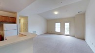 This single-floor residence is designed with an open and functional layout that connects the living area to the kitchen seamlessly. The spacious living room features vaulted ceilings, neutral-toned carpet, and large windows alongside a glass-paneled exterior door that allows natural light to fill the space and provides outdoor access. A curved kitchen peninsula serves as a natural divider between the living and kitchen areas, offering additional counter space and a casual dining option.

The kitchen is well-appointed with warm-toned wood cabinetry extending to the ceiling, ample upper and lower storage, light-colored countertops, and white appliances including an electric range with overhead microwave and a full-size refrigerator. Tile flooring grounds the kitchen area, contrasting with the carpeted living spaces.

The bedrooms are comfortably sized, each featuring plush carpet, neutral walls, and windows with blinds. At least one bedroom includes a ceiling fan with light fixture, enhancing ventilation and comfort. An additional smaller room offers flexible use as a bedroom, office, or storage space.

The bathrooms are finished with classic patterned tile flooring, wood-toned vanities with light countertops, mirrors, and full tub-shower combinations, providing practical and clean design throughout.