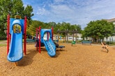 Located at 8160 Veterans Pkwy in Columbus, Georgia, this well-appointed outdoor community space serves as a central gathering hub within a residential community. The grounds are thoughtfully landscaped with mature trees that provide generous natural shade, complemented by manicured shrubs and wood chip ground cover throughout the activity areas.

At the heart of the space is a vibrant playground equipped with colorful play structures, including slides and climbing elements, set on a safe wood chip surface. Seating benches are conveniently positioned nearby, allowing residents to supervise children or simply enjoy the outdoors. A rustic wooden pergola adds architectural character to the space, offering a shaded retreat alongside additional seating benches.

The community also features a dedicated picnic area with sturdy outdoor tables, a paved patio surface with decorative stonework, and an accessible grill station — ideal for casual outdoor dining and social gatherings. Curving concrete walkways connect the various amenity zones, creating a cohesive and easy-to-navigate layout.

The surrounding multi-story residential buildings, constructed with a combination of stone and siding exteriors, frame the space and contribute to an inviting neighborhood atmosphere. Open grassy areas provide additional room for recreation, while the overall design encourages an active and connected community lifestyle under the open Georgia sky.