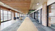 This single-floor commercial office space masterfully combines industrial character with contemporary design across a generously proportioned floor plate. The open-plan main area serves as the centerpiece, featuring a striking slatted wood ceiling with exposed structural elements, original brick walls, and expansive steel-framed windows that flood the space with natural light and offer outward views of the surrounding waterfront and urban environment. Freestanding workstations are arranged throughout this area, promoting a collaborative and open working atmosphere.

A central corridor, finished with a light-toned resilient floor strip flanked by grey modular carpet tiles, connects the open workspace to a series of enclosed private offices. These offices are accessed through sleek black-framed glass partition walls, maintaining visual connectivity while offering acoustic separation. Each private office features a drop ceiling with recessed lighting, soft blue accent walls, and grey carpet tile flooring, creating a calm and focused environment suitable for individual or small-group work.

Throughout the space, exposed green mechanical ductwork and concrete ceiling elements in the transitional zones add an honest industrial aesthetic. The combination of warm wood tones, exposed brick columns, and modern glass partitions gives the space a refined yet approachable character, well-suited for a professional office environment.