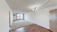 This single-floor unit offers three bedrooms, one bathroom, a kitchen, a hallway, and two closets within a compact yet functional layout. The living area is the heart of the home, featuring a combination of rich hardwood-style flooring and plush carpeting, a textured ceiling, and an elegant chandelier. A large sliding glass door floods the space with natural light and provides access to a balcony with outdoor views, while a wall-mounted AC unit ensures year-round comfort. The galley-style kitchen is well-equipped with warm wood cabinetry offering ample upper and lower storage, white appliances including a range and refrigerator, a double-basin sink, and light countertops — all set on hardwood-style flooring beneath a textured ceiling. The generously sized primary bedroom is carpeted and features a wide window that frames outdoor views of surrounding trees, along with baseboard heating. The additional bedrooms are similarly carpeted and naturally lit. The full bathroom includes a wood-framed vanity with a mirrored medicine cabinet, a tiled tub/shower combination, mosaic tile flooring, and warm vanity lighting. Two closets provide practical storage throughout the unit.