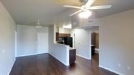 This single-story space features an open-concept design that seamlessly connects the living area and kitchen. Dark hardwood flooring runs throughout, providing a rich contrast against the light gray walls and white trim. The living area is equipped with a ceiling fan with integrated lighting, contributing to the overall brightness of the space. A peninsula-style kitchen counter with granite countertops serves as a natural divider between the living and kitchen areas. The kitchen features dark wood cabinetry, black appliances including a refrigerator and microwave, and an under-cabinet light fixture that illuminates the workspace. A hallway visible from the main living area leads to what appears to be a bathroom, which includes a bathtub and additional cabinetry. The front entry door is positioned along the left wall, framed by white molding that carries throughout the space. HVAC vents are integrated into the ceiling, ensuring climate comfort. The layout feels functional and well-organized, with clear sightlines across the entire living space from any vantage point.