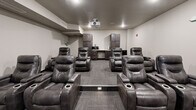 Located at 704 First Dr NW in Austin, Minnesota, this purpose-built theater room offers a fully immersive cinematic experience within a compact, efficiently designed single-floor layout. The room is configured with multiple rows of plush, dark leather power recliner seats arranged in a tiered formation, each equipped with built-in cup holders and wide armrests for maximum comfort during extended viewing sessions. A ceiling-mounted projector delivers crisp imagery onto a large wall-mounted projection screen positioned at the front of the room, ensuring clear sightlines from every seat. The neutral gray walls and low ceiling with recessed lighting create a dark, atmosphere-enhancing environment ideal for an immersive viewing experience. Durable, patterned carpet tiles in a neutral gray tone cover the flooring throughout, providing both practicality and a polished aesthetic. At the rear of the room, dark wood cabinetry flanks a built-in refreshment counter complete with a microwave and mini-refrigerator, allowing guests to enjoy snacks and beverages without leaving the space. A subwoofer is also present, contributing to the room's audio capabilities. Safety features include a visible fire alarm system. A single entry door provides a clean, controlled access point, completing this well-appointed, self-contained theater experience.
